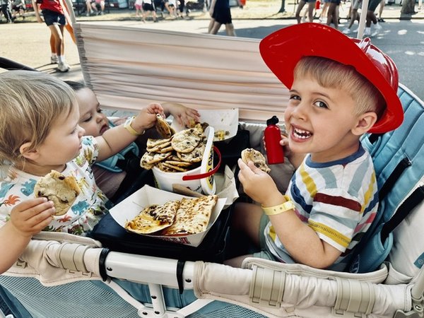 Two young children eating at a fair, one wearing a red firefighter hat